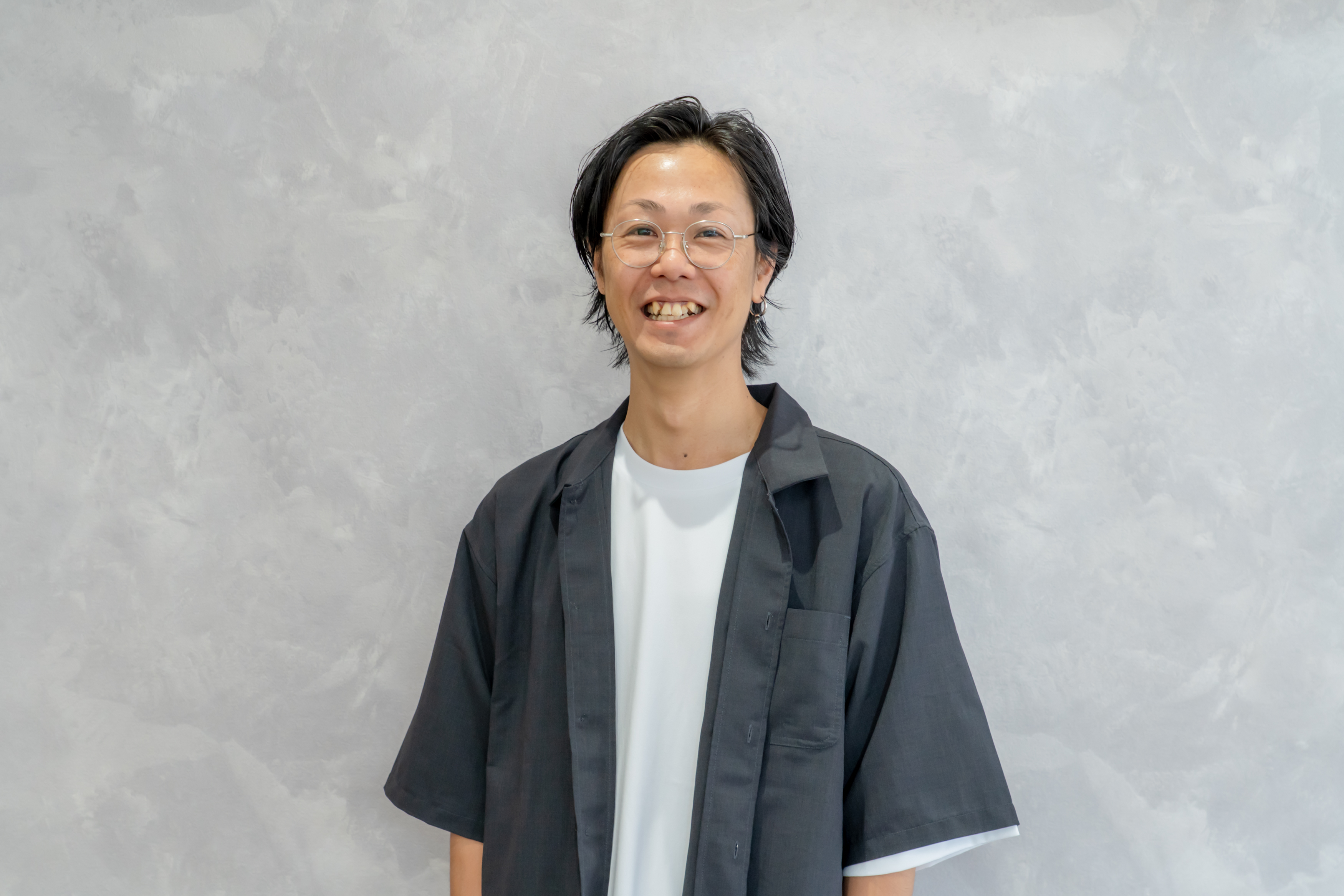 落ち着いた個室サロンで自然な笑顔を見せる男性スタイリスト、黒のトップスで清潔感のあるポートレート